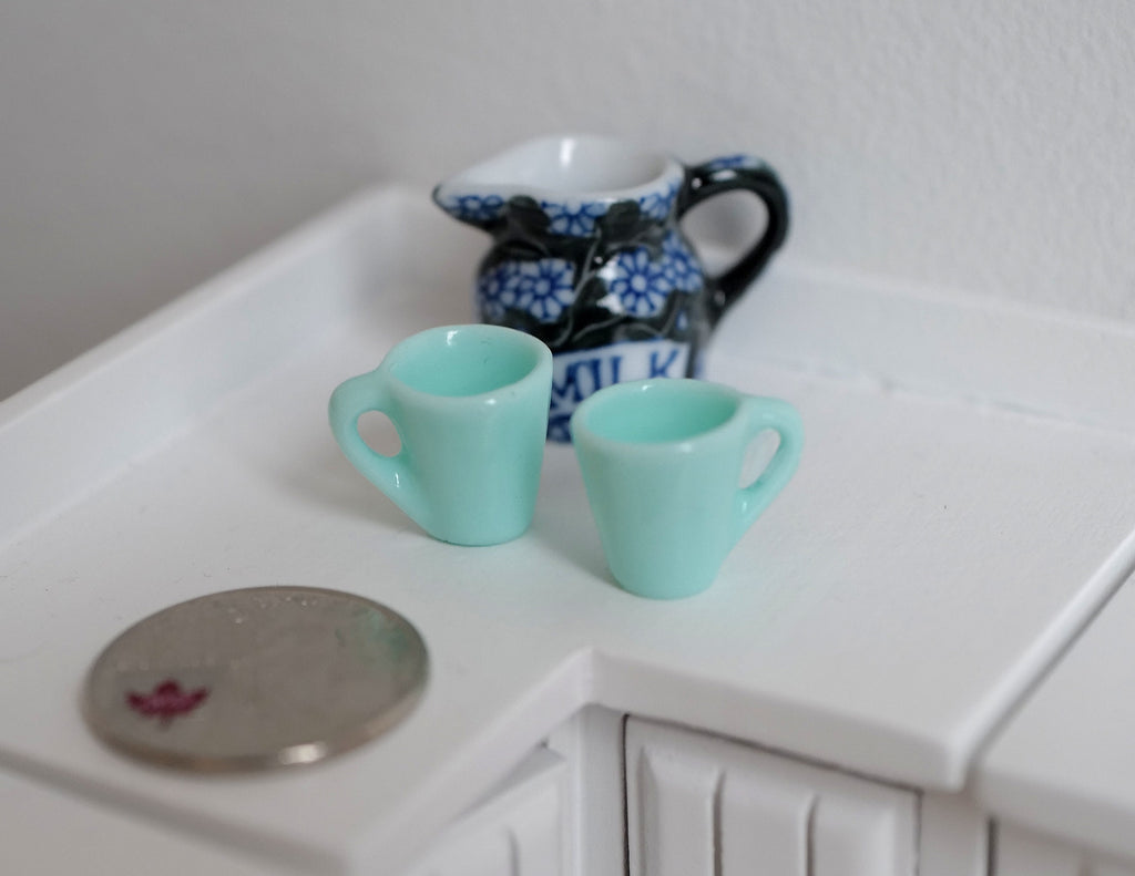 1:12 scale miniature tea cups in mint green, set of 2, shown on a white kitchen counter beside a coin and a blue jug