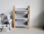1:12 scale miniature wood open storage shelf with 3 gray shelves and natural wood posts, shown beside an armchair