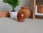 1:12 scale miniature clay round jar pot in glossy brown, shown beside terracotta pots and a potted plant