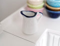 1:12 scale miniature ceramic milk jug in enamelware style, white with navy blue rim, on a white counter by bowls