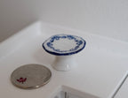 1:12 scale miniature white pedestal cake stand with blue pattern rim, shown on a white counter beside a coin for scale