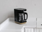 1:12 scale miniature black drip coffee maker with clear glass carafe, shown on a white kitchen counter