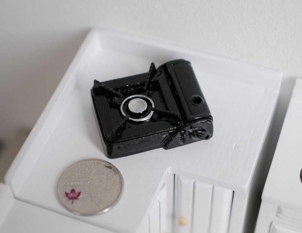 1:10 scale miniature black portable gas stove with a single burner, shown on a white counter beside a coin