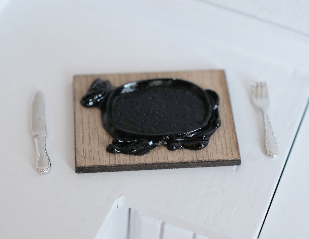 1:12 scale miniature cow shaped black sizzler steak plate with wood board, fork and knife, shown on a white table