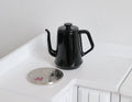 1:12 scale miniature metal gooseneck kettle in black with lid, shown beside a coin on a white counter