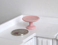 1:12 scale miniature ceramic pedestal cake stand in pink, shown on a white kitchen counter beside a coin for scale