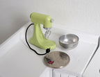 1:12 scale miniature stand mixer in green with silver bowl and black cord, shown on a white counter beside a coin