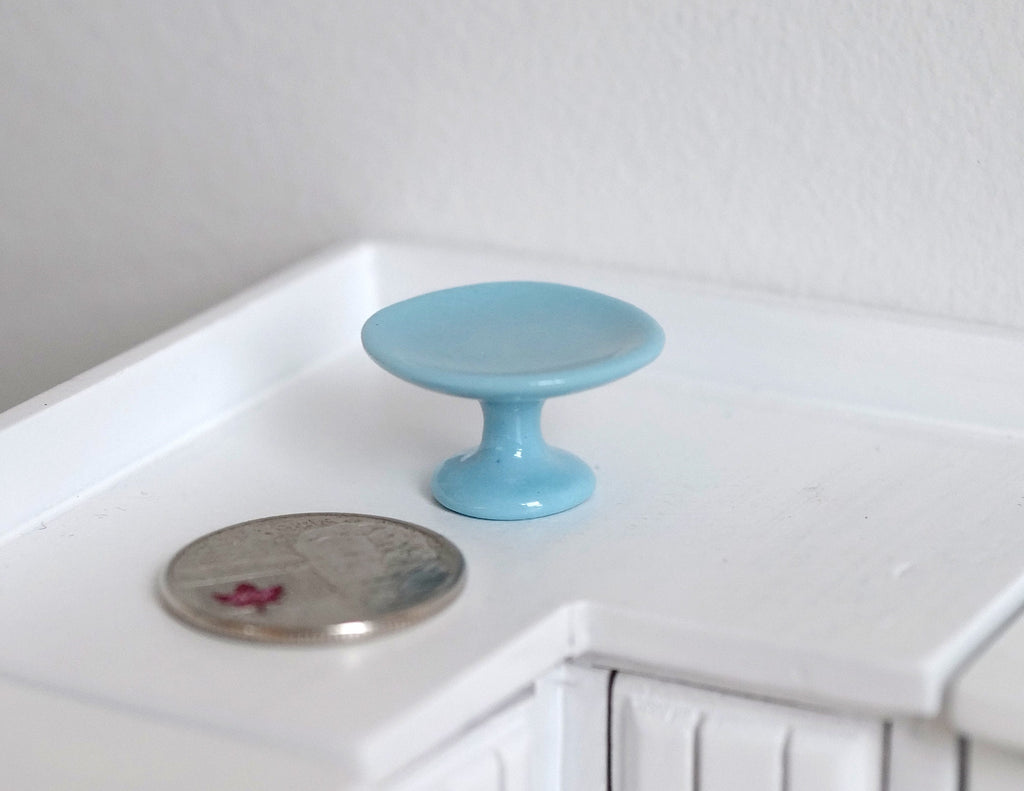1:12 scale miniature blue ceramic pedestal cake stand, shown on a white kitchen counter beside a coin for scale
