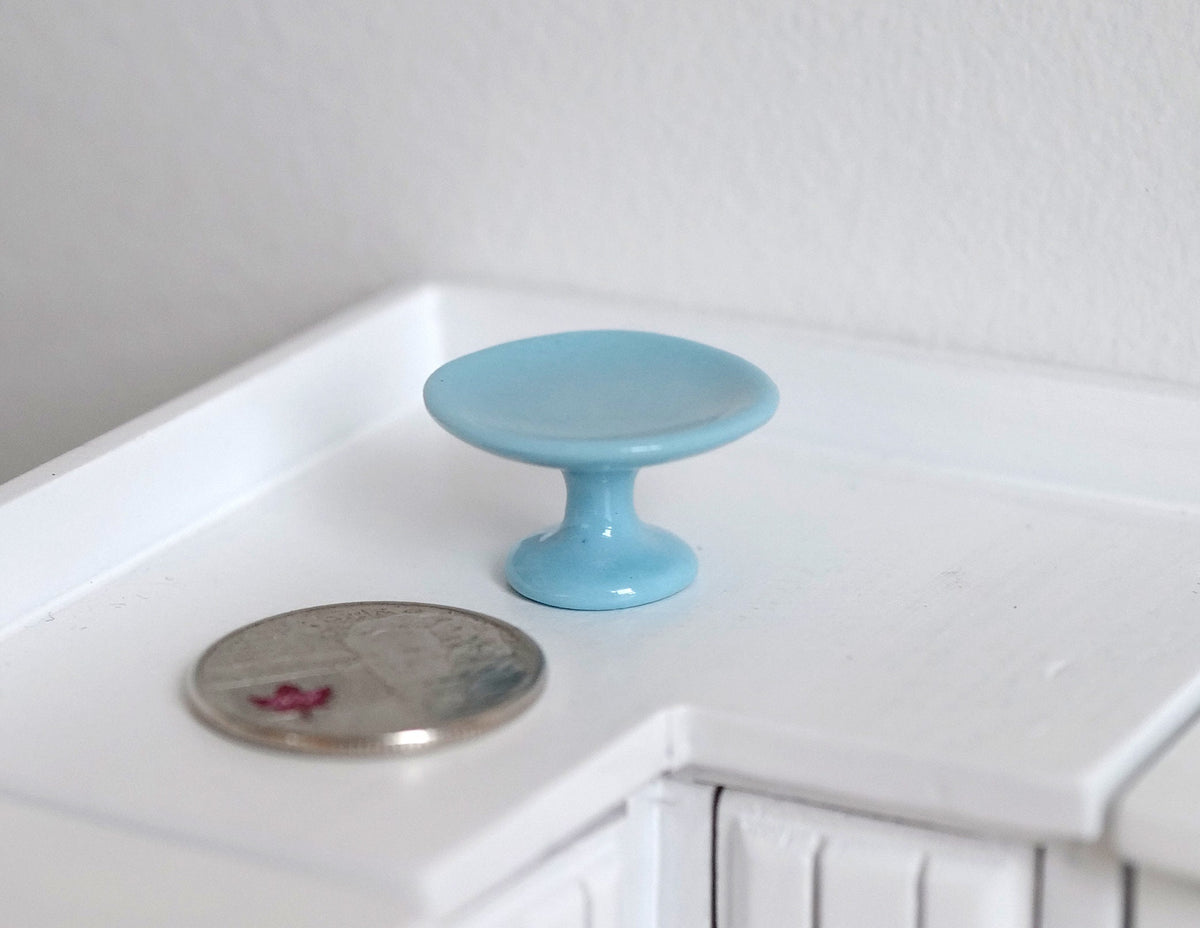 1:12 scale miniature blue ceramic pedestal cake stand, shown on a white kitchen counter beside a coin for scale