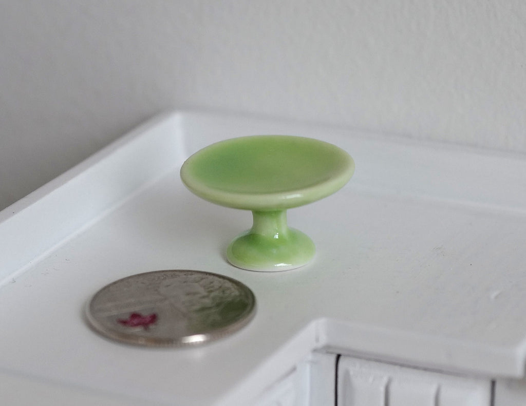 1:12 scale miniature ceramic pedestal cake stand in green, shown beside a coin on a white kitchen counter