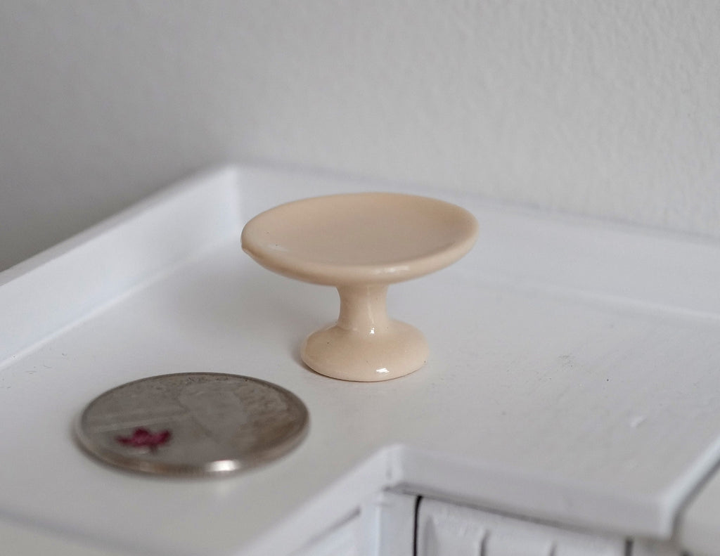 1:12 scale miniature ceramic pedestal cake stand in beige, shown on a white kitchen counter beside a coin for scale