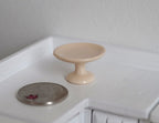 1:12 scale miniature ceramic pedestal cake stand in beige, shown on a white kitchen counter beside a coin for scale