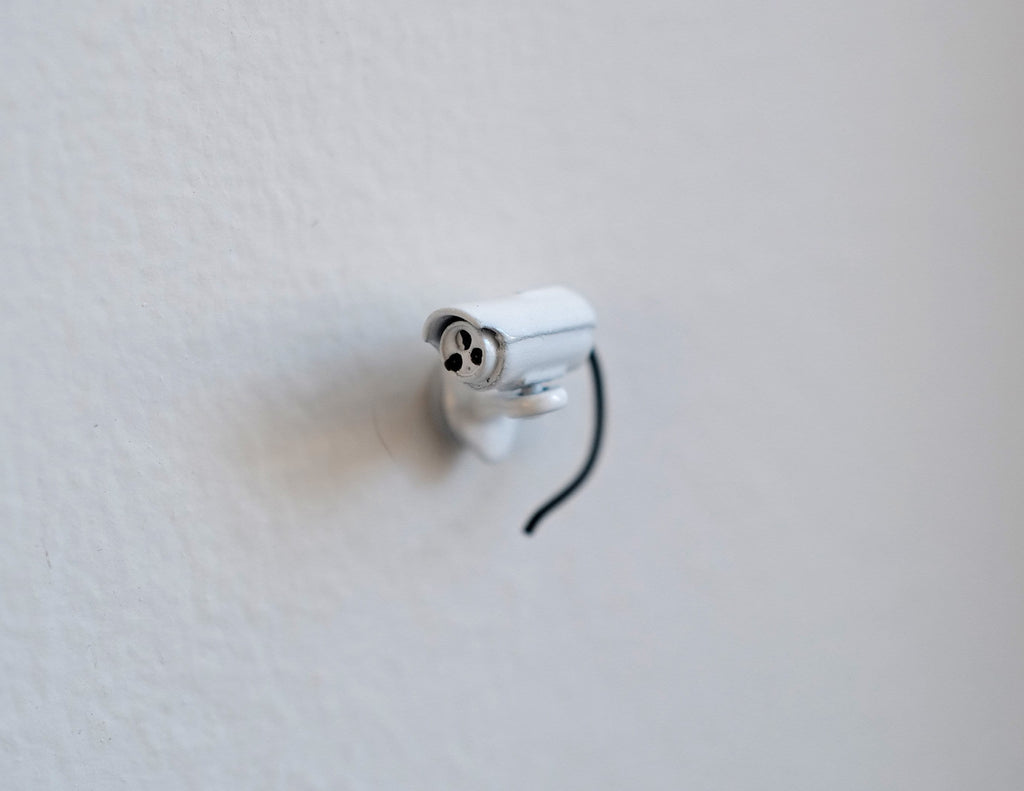 1:12 scale miniature bullet security camera in white with antenna and black cable, shown mounted on a white wall