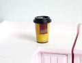 1:6 scale miniature coffee cup in yellow with red sleeve and black lid, shown on a white counter
