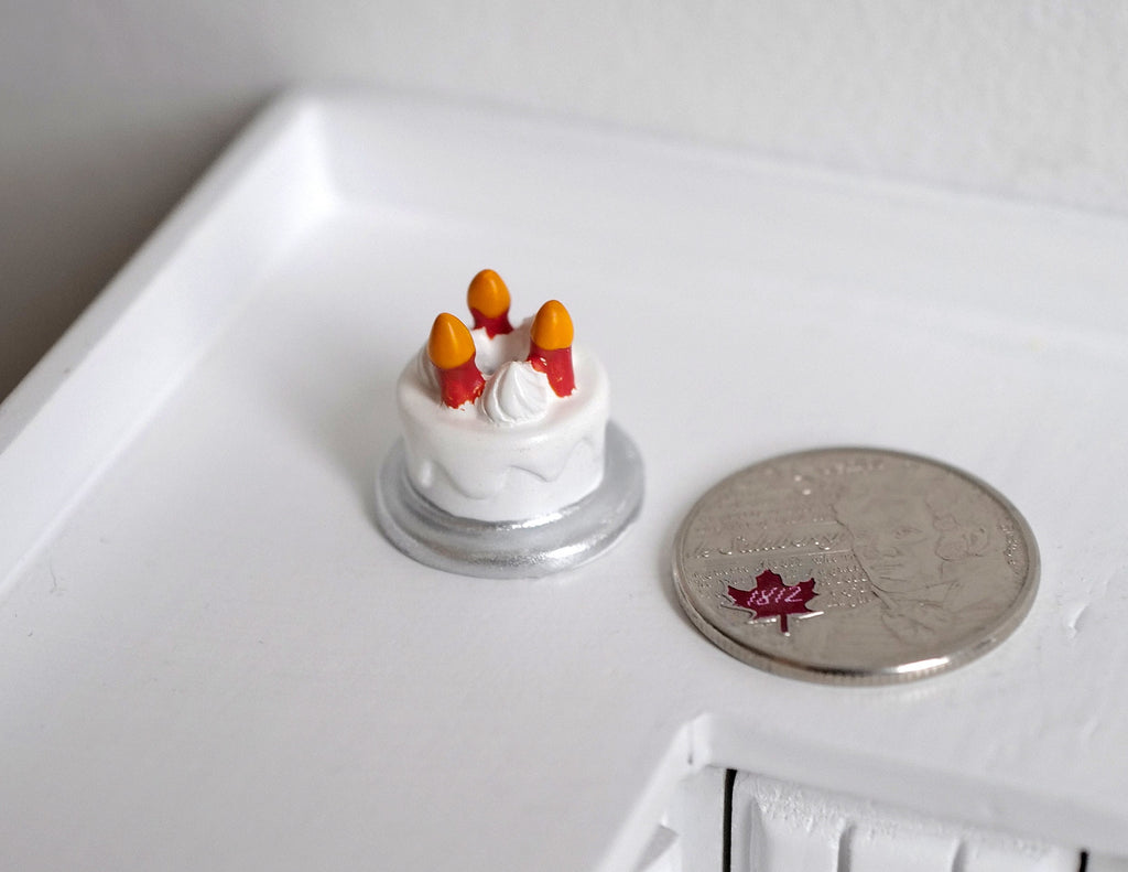 1:12 scale miniature Christmas cake with white icing and orange fruit on a silver board, beside a coin for scale