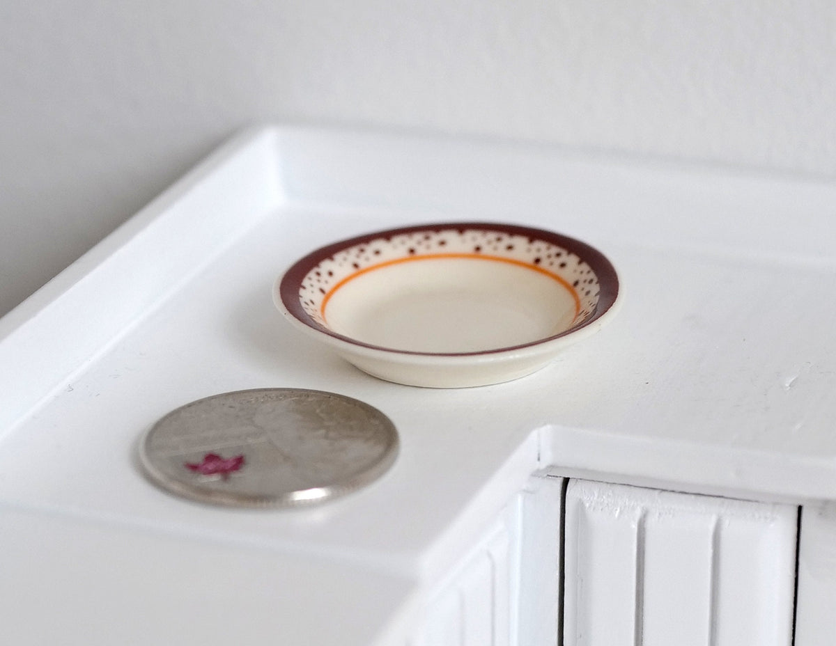 1:12 scale miniature cream ceramic shallow bowl with red patterned rim, shown on a white counter beside a coin