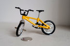 1:12 scale miniature kid size freestyle bike in yellow with front and rear pegs and black tires, beside a coin
