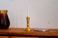1:12 scale miniature gold candlestick holder with white candle and round base, shown on a wood shelf beside a coin