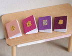 1:12 scale miniature books that open, pastel covers with gold emblems, set of 4 in pink and purple, on a wood table