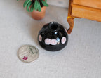 1:12 scale miniature black ceramic squat vase with white and pink polka dots, shown on a floor beside a coin for scale