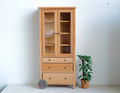 1:12 scale miniature wood tall kitchen cabinet in natural finish with glass doors, 4 shelves and 3 drawers, by a plant