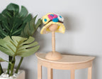 1:12 scale miniature cloche hat with floral patchwork in yellow, pink and blue on a wood hat stand, on a side table