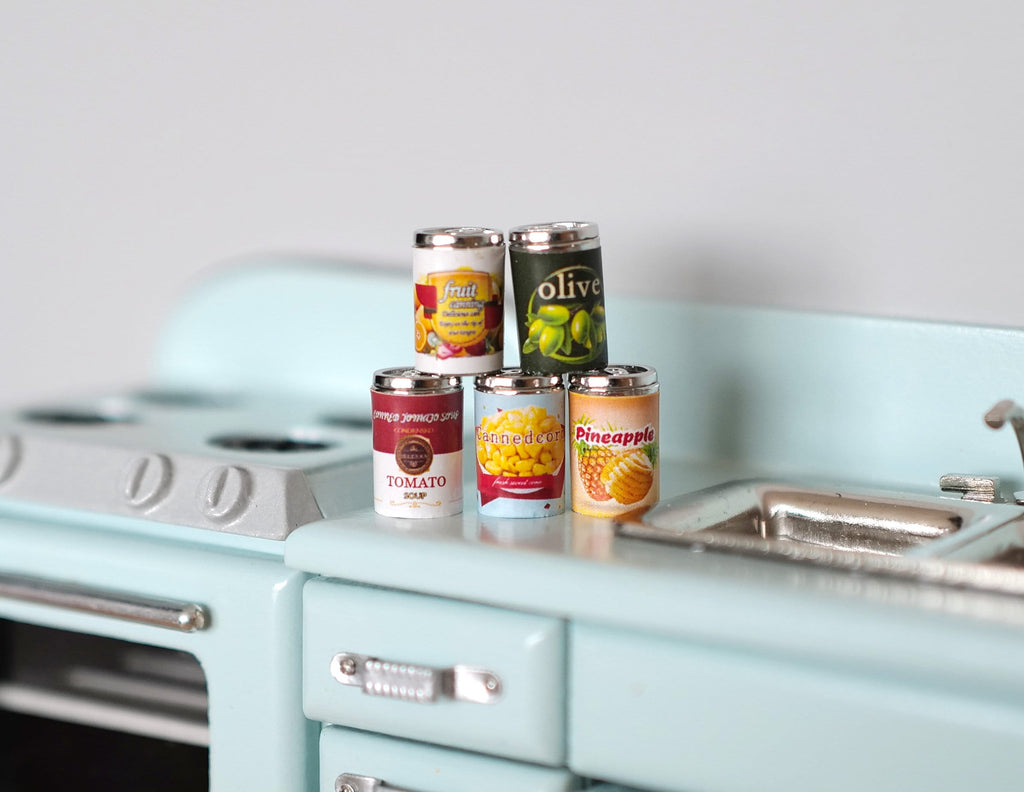 1:12 scale miniature metal fruit tin cans with printed labels, set of 5, stacked on a pale blue kitchen counter