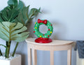 1:12 scale miniature decorated Christmas wreath on a red stand, green with red bow and ornaments, on a side table