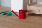 1:12 scale miniature metal watering can in red with spray nozzle, shown on a wood surface with a green hose coil