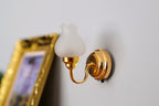 1:12 scale miniature battery LED wall light in brass with frosted white shade and on off switch, on a gray wall