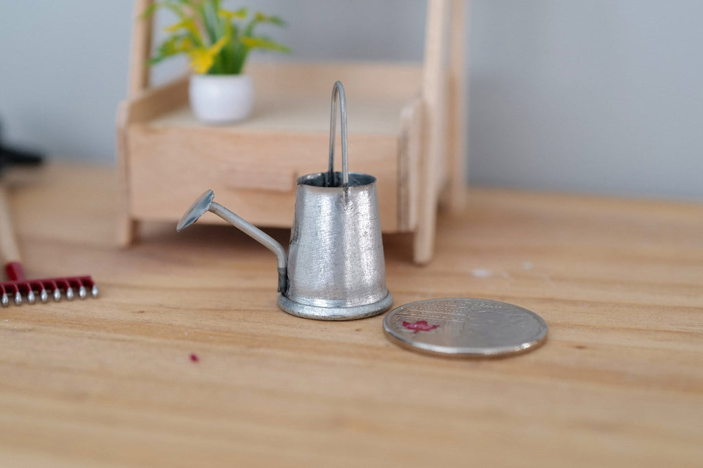 1:12 scale miniature galvanized metal watering can with long spout, silver gray, on a wood floor beside a coin for scale