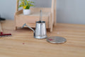 1:12 scale miniature galvanized metal watering can with long spout, silver gray, on a wood floor beside a coin for scale