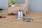 1:12 scale miniature galvanized metal watering can with long spout, silver gray, on a wood floor beside a coin for scale
