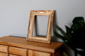 1:12 scale miniature ornate rectangular picture frame in champagne gold, shown empty leaning on a wood dresser