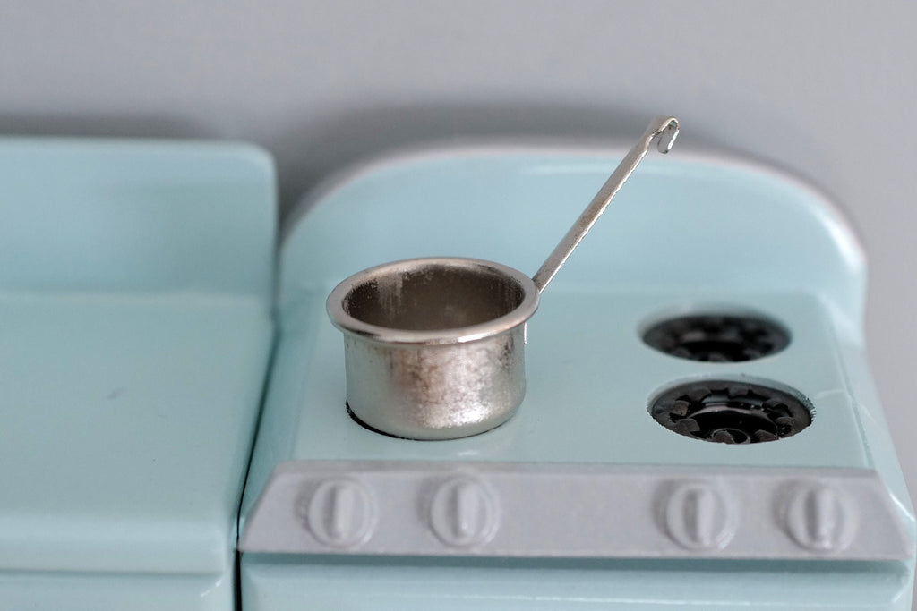 1:12 scale miniature silver metal hanging saucepan with long handle, shown on a mint green stove