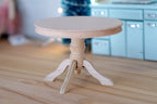 1:12 scale miniature unpainted wood round pedestal dining table with 3 curved feet, shown on a wood floor