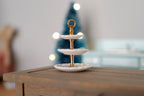 1:12 scale miniature ceramic 3 tier cake stand in white and gold with a ring handle, shown on a wood table
