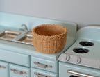 1:12 scale miniature round woven basket in natural color, shown on a light blue kitchen counter beside a sink