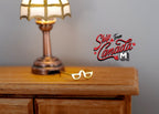 1:12 scale miniature metal eyeglasses in gold, shown on a wood dresser beside a lit table lamp