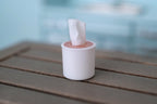 1:12 scale miniature tabletop tissue roll holder in white and pink with tissue pulled up, shown on a wood table