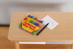 1:12 scale miniature wax crayons in a yellow box with colorful crayon print, shown on a wood desk with paper and pencil