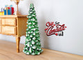 1:12 scale miniature snow covered Christmas tree, green with white flocked tips, shown on a floor beside a wood desk