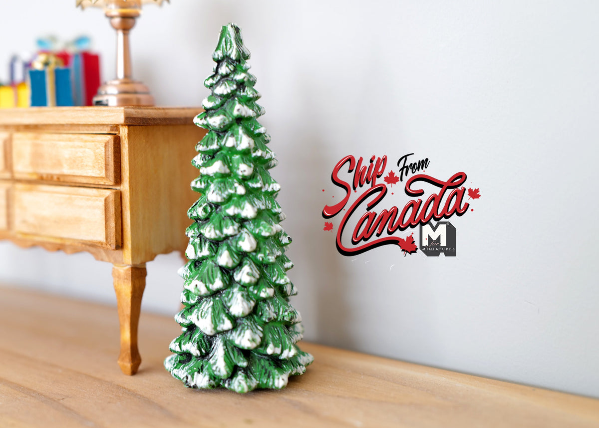 1:12 scale miniature snow covered Christmas tree, green with white flocked tips, shown on a floor beside a wood desk