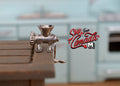 1:12 scale miniature metal meat grinder in silver with hand crank and table clamp, clamped to a wood table