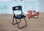 1:12 scale miniature metal folding chair in black with a cutout back, shown open on a wood floor