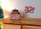 1:12 scale miniature ceramic teapot with lid in pink with floral decoration, shown on a wood dresser