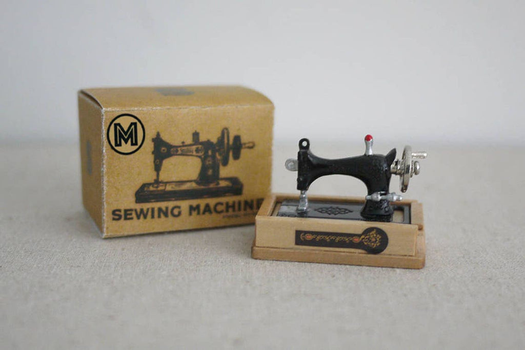 1:12 scale miniature classic sewing machine in black on a wood base, shown beside its printed kraft packing box