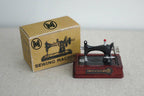 1:12 scale miniature sewing machine with box and notions, black machine on a red wood base beside its printed box