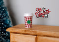 1:6 scale miniature coffee cup with red and white gingham sleeve and white lid, shown on a wood dresser by a tree