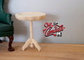 1:12 scale miniature unpainted wood round pedestal end table with tripod base and turned column, shown beside a green armchair